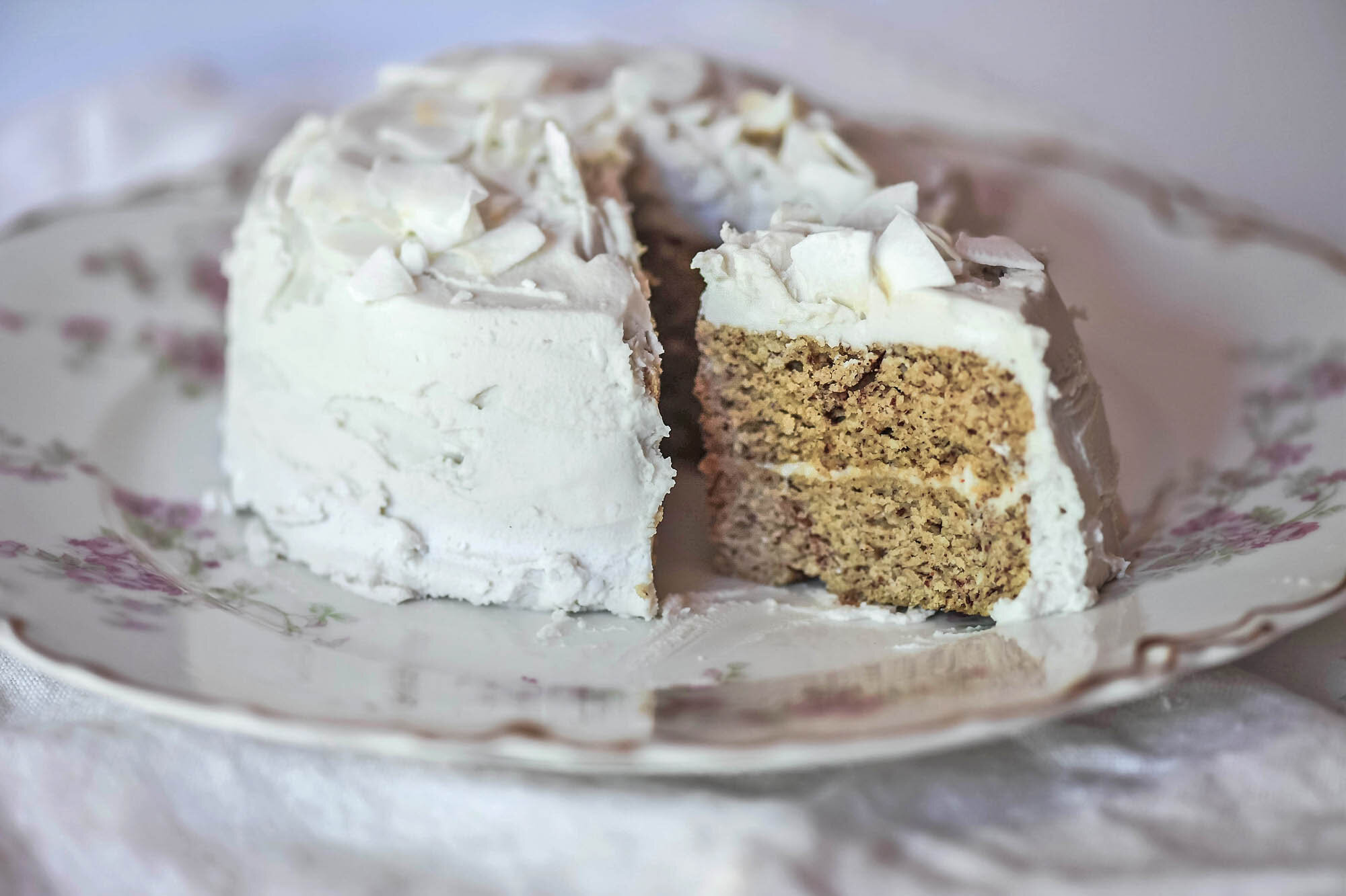 Slice removed from mini gluten free coconut cake topped with coconut flakes on decorative floral plate.