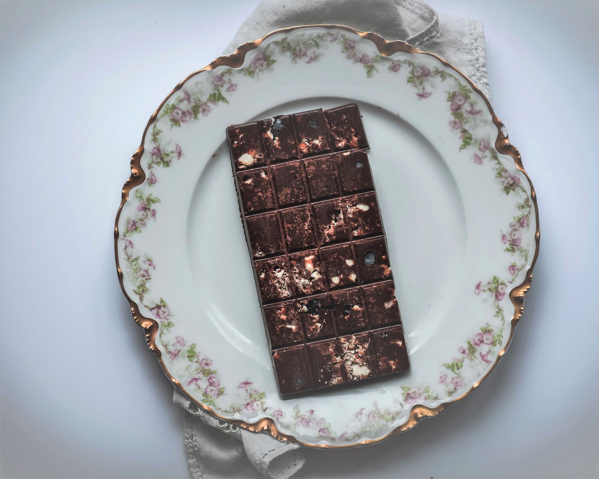 Homemade chocolate bar with chopped almonds and freeze dried blueberries atop decorative plate.